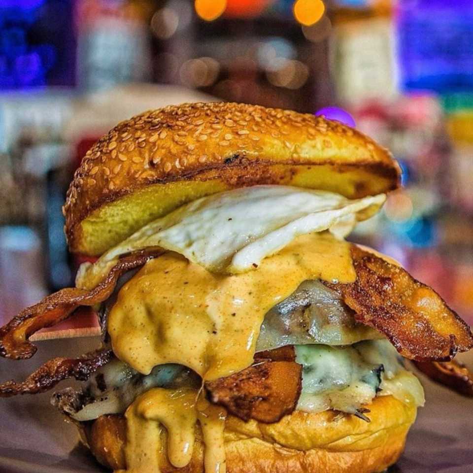 Stacked burger with bacon, melted cheese, sauce, and a fried egg on a sesame bun.
