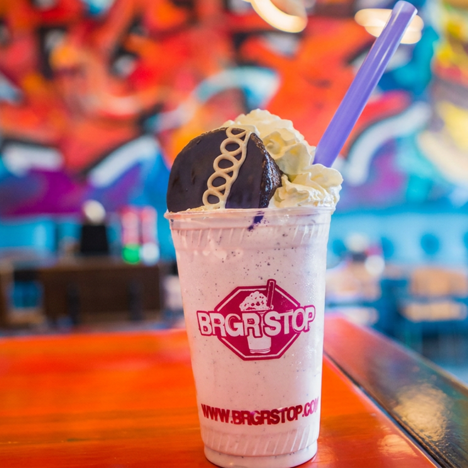 Pink milkshake topped with whipped cream and chocolate cookie, served in a branded Burgerstop cup with a purple straw.
