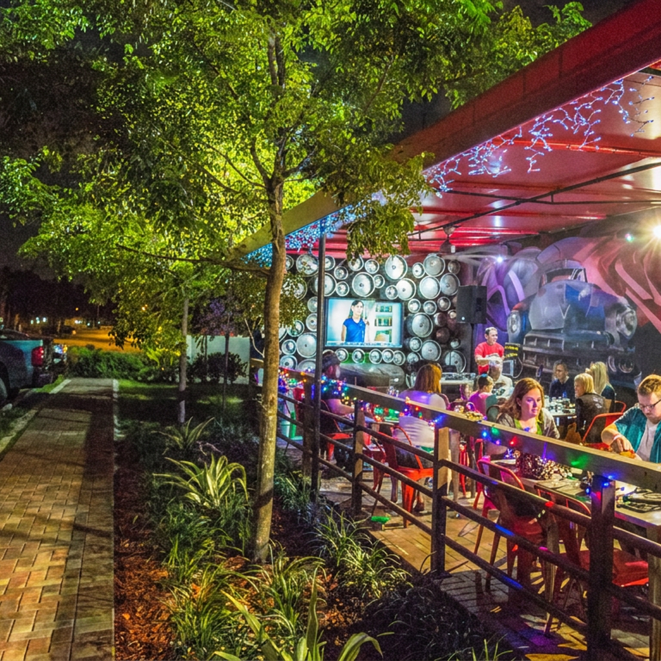 Nighttime view of a lively restaurant patio with diners, colorful lights, murals, televisions, and landscaped walkway.