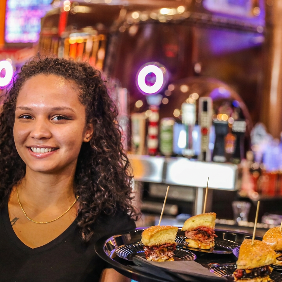 Smiling server holds a tray of four miniature burgers in a lively, colorful restaurant bar.