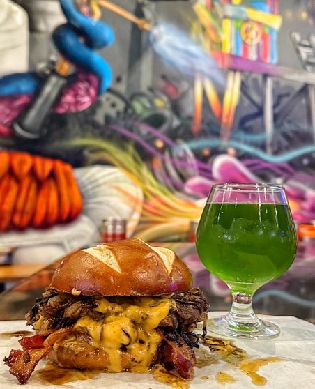 IThe Bavarian Beermeister burger at Brgr Stop — two Worcestershire-infused smash burgers layered with onions, beer cheese, applewood bacon, and sweet Bavarian mustard on a toasted pretzel bun, served with a glass of green beer.